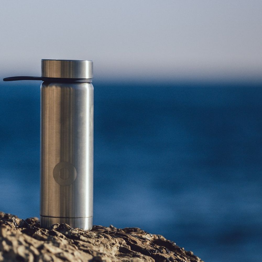 Slim silver metal water bottle standing on a rock with the sea behind it